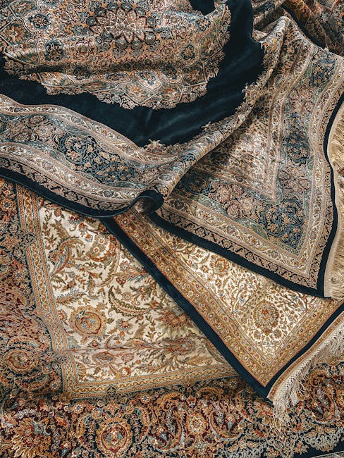 Close-up image of intricately patterned Persian-style rugs with detailed floral and geometric motifs in shades of beige, brown, blue, and gold, layered on top of each other on a hardwood floor. The ru