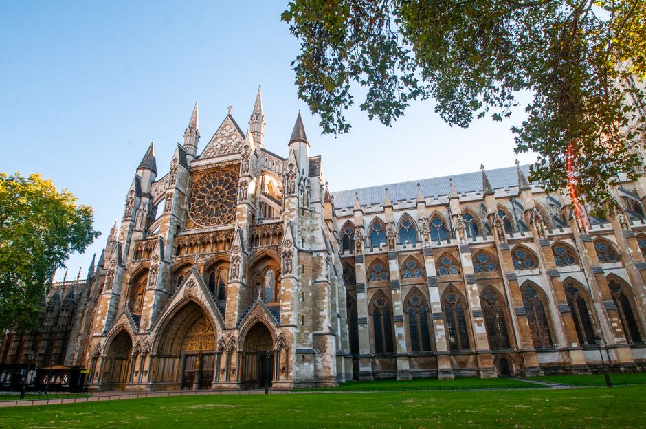 The image shows the exterior of Westminster Abbey, featuring its elaborate gothic architecture with tall, pointed spires and detailed stone carvings. The facade is constructed from light-colored stone